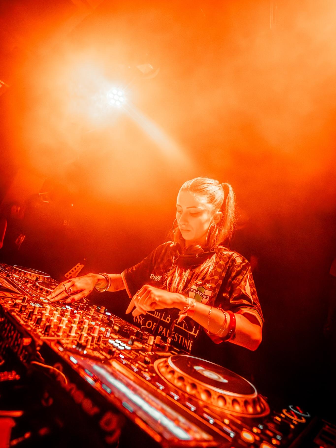 A DJ mixing under orange haze and a single beam of light at an OLYMPE night